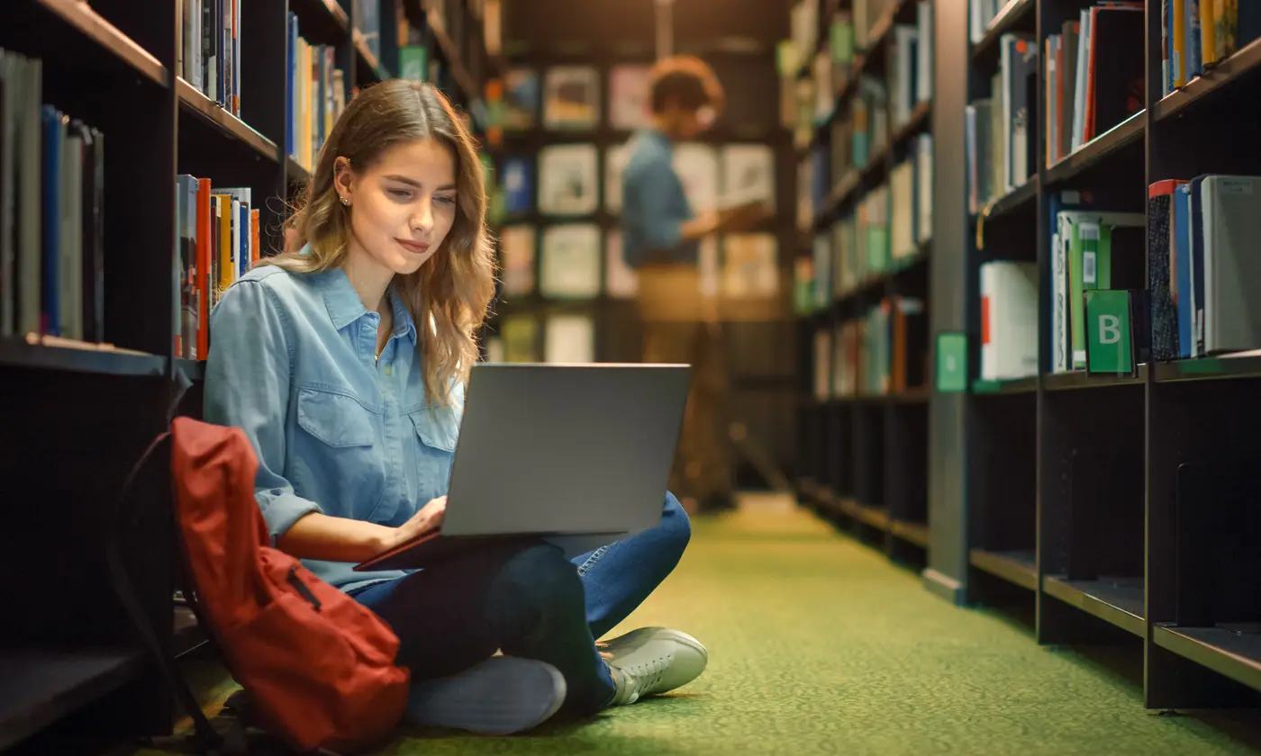 Girl siting on floor using laptop in library aisle